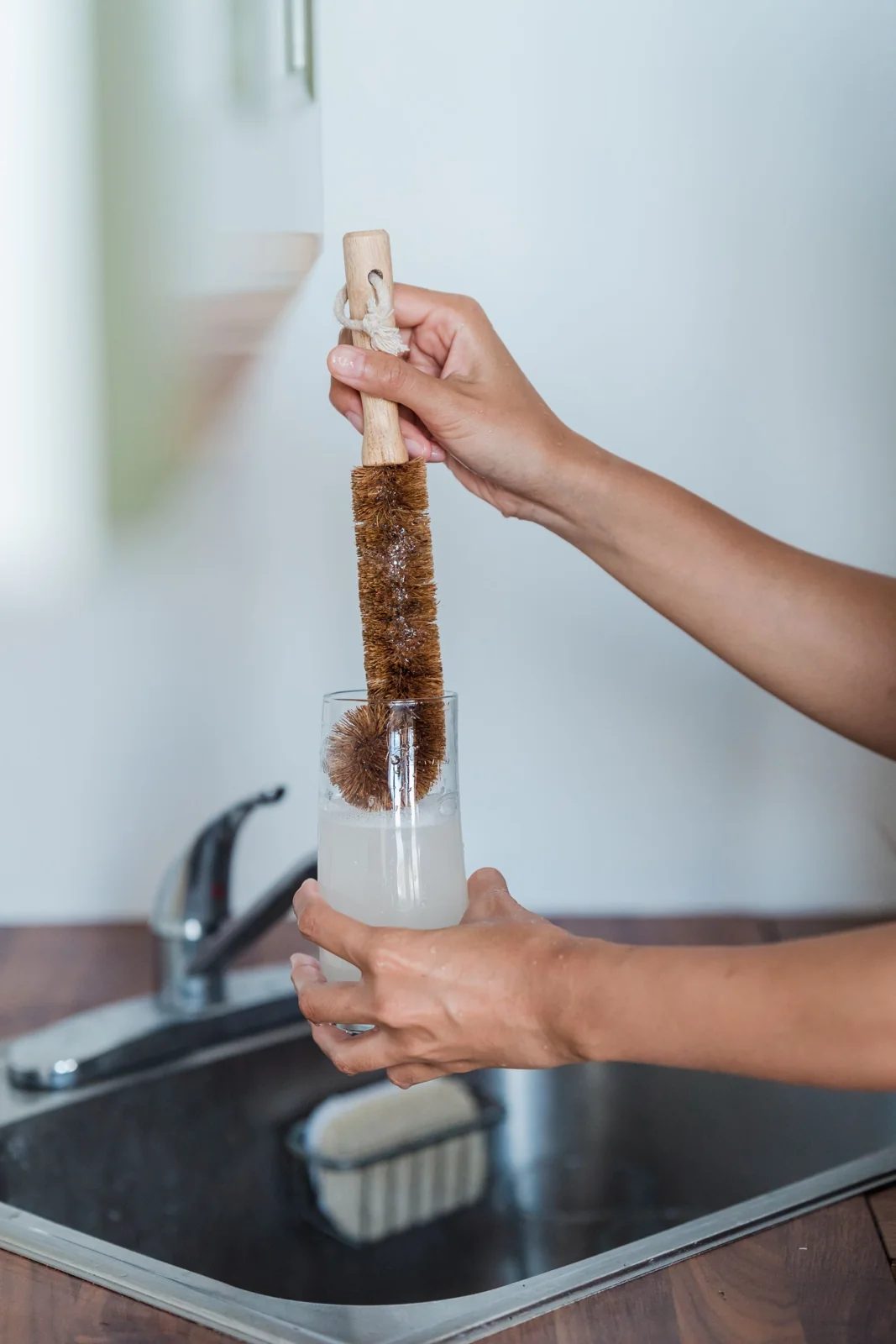 Hands cleaning glassware with a natural brush at a sink